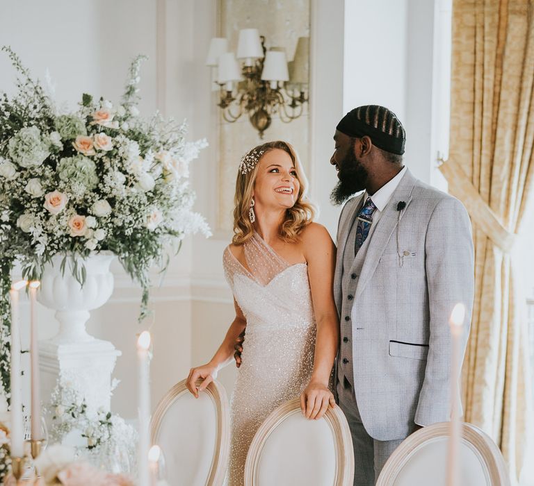 Bride in a sparkly wedding dress with side swept curly hair smiling at her groom in a light grey suit at their elegant wedding reception 