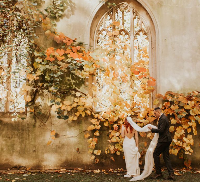 Groom holds his brides veil for Autumnal wedding day photography with yellow and orange leaves,