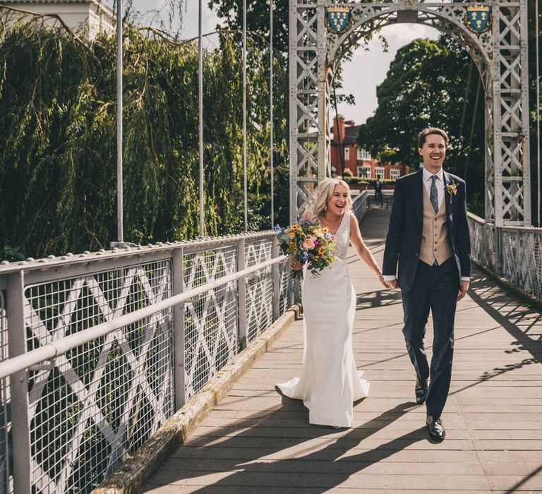 Laughing bride in Justin Alexander wedding dress and colourful bouquet holds hands with laughing groom on bridge in Chester