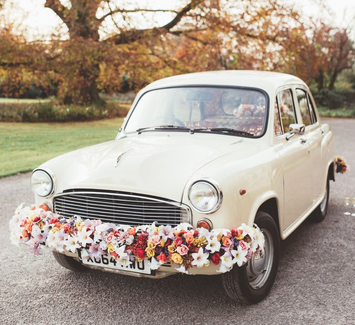 A cream vintage car with a bright multicoloured  flower garland running along the front bumper.