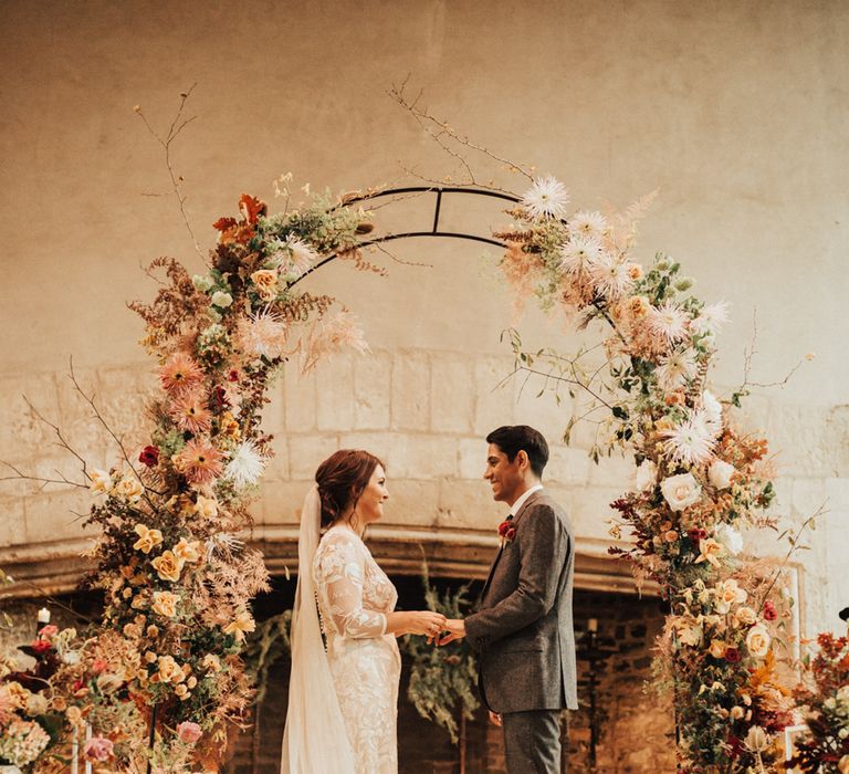 Bride in a lace wedding dress with long sleeves holding her grooms hand at the altar in a world suit surrounded by autumn wedding flowers and pillar candles 