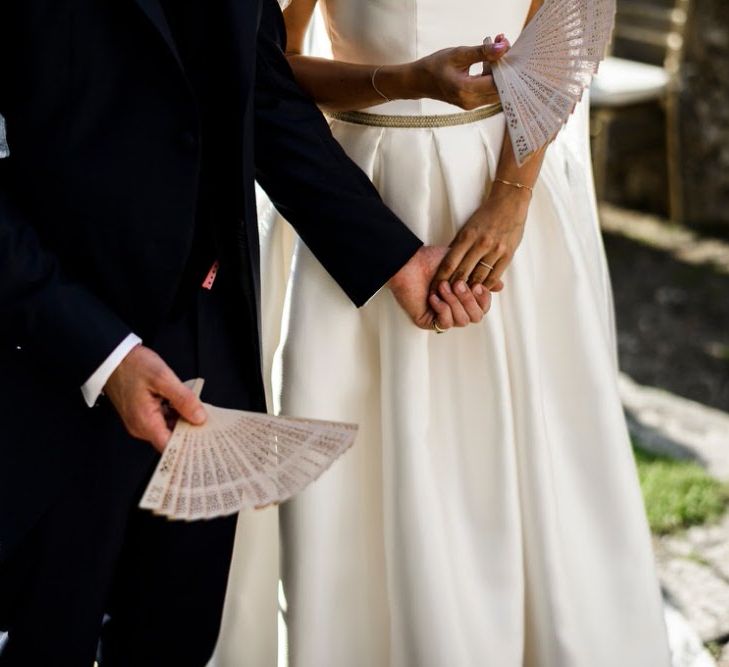 Closeup of bride and Groom holding hands with groom also holding a white fan - Image courtesy of Marble Private