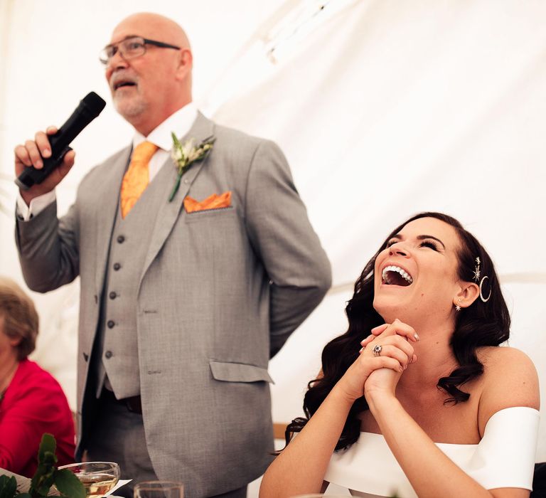 Laughing bride with long curled hair and celestial hair pins clasps her hands sat at top table whilst father in grey Hugo Boss suit with waistcoat and yellow tie talks into microphone