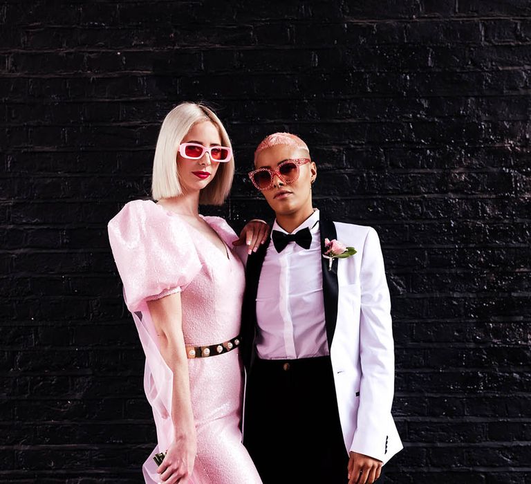 Alternative wedding fashion with bride in a pink jumpsuit and sunglasses and bride in a white tuxedo jacket with black lapel and bow tie 