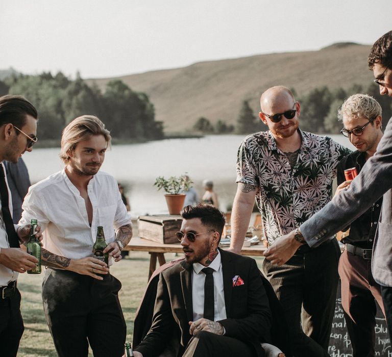 Groom with tattoos wearing a black suit and loafers, sitting with his groomsmen at moody romantic wedding