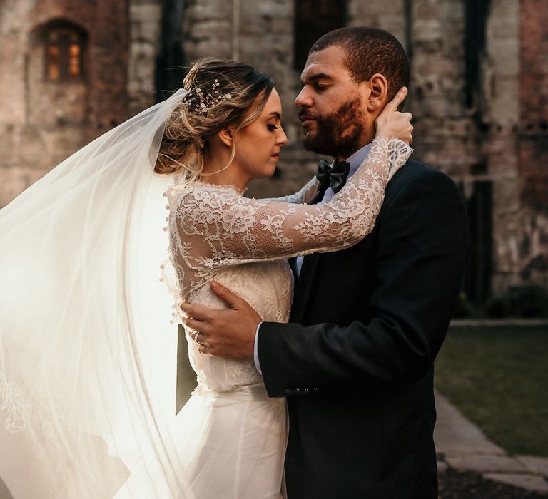 Bride and groom portrait with bride in a lace long sleeve wedding dress holding her grooms head as her appliqué veil blows in the wind 