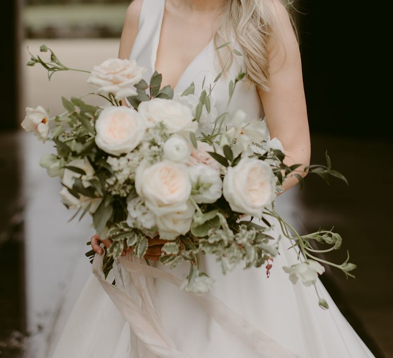 Bride wears her blonde hair in loose and natural curls with white bridal headband, holding white floral bouquet 