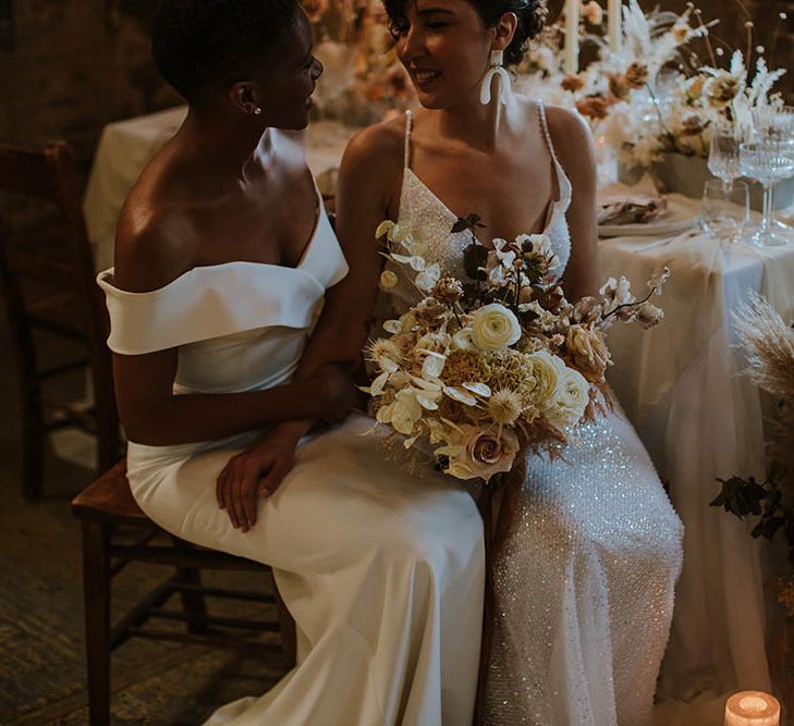 Bride in an off the shoulder wedding dress sitting with her bride in a sparkly wedding dress holding a cream and beige neutral wedding bouquet 