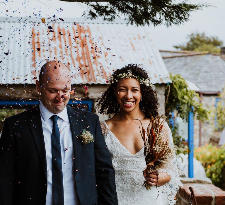 Bride & groom walk along together whilst holding hands as bride wears lace wedding gown and holds dried floral bouquet for Cornwall elopement