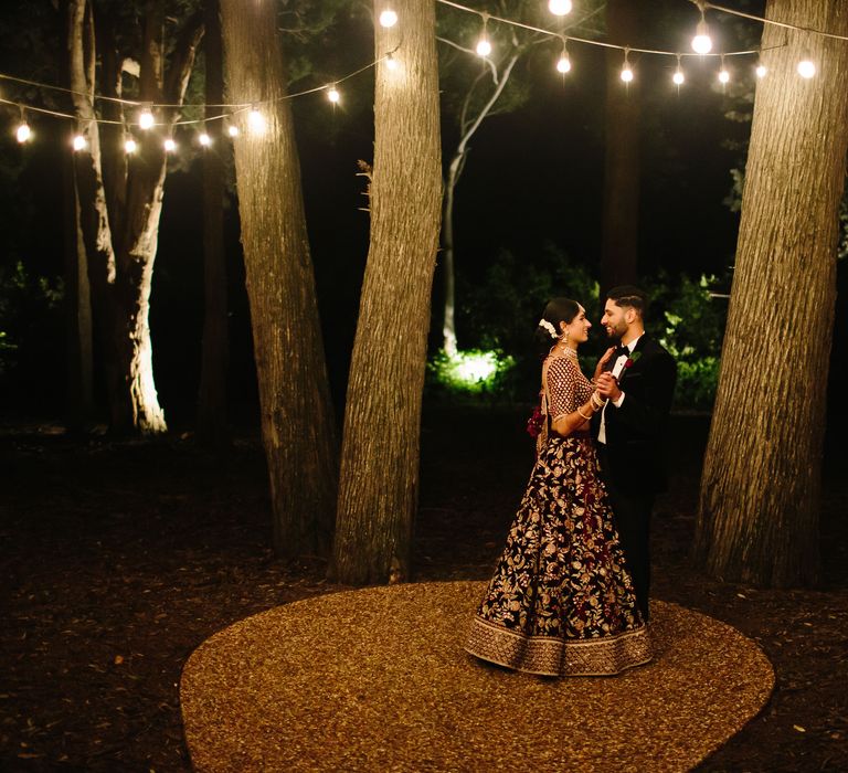 Bride & groom embrace outdoors as fairy lights hang from trees outdoors during the night