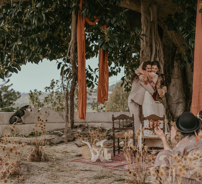 Bride & groom hug one another under tree outdoors in Sicily after wedding ceremony as guests clap