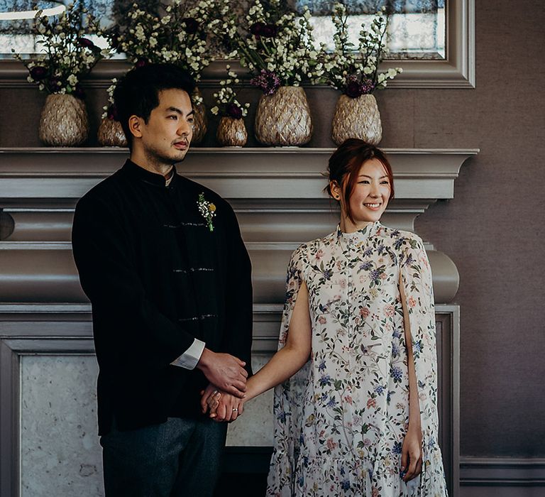 Intimate Old Marylebone Town hall wedding ceremony with East Asian groom in a navy velvet jacket holding his bride's hand in a short blue floral wedding dress 