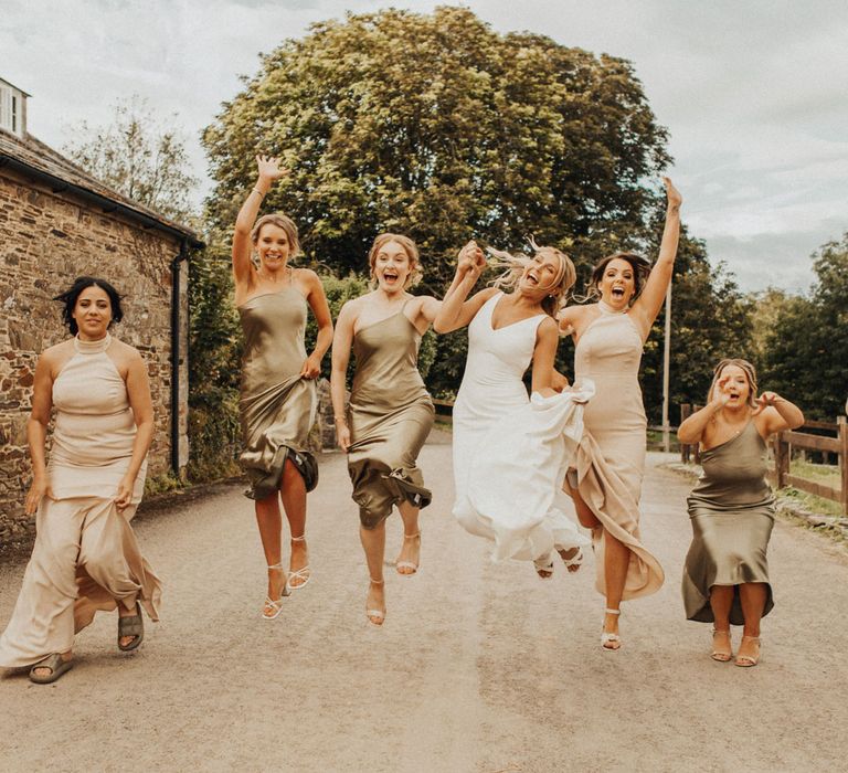 Bride in white Made With Love wedding dress and bridesmaids in sage green and beige bridesmaids dresses jump in the air on country lane at Anran Devon