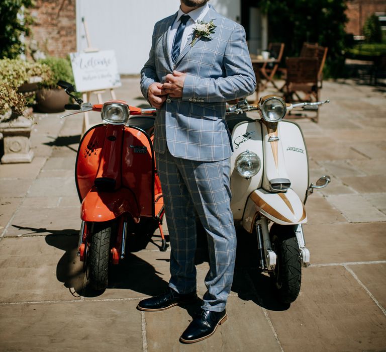 Groom in a pale grey gingham check suit standing next to his vespa scooter