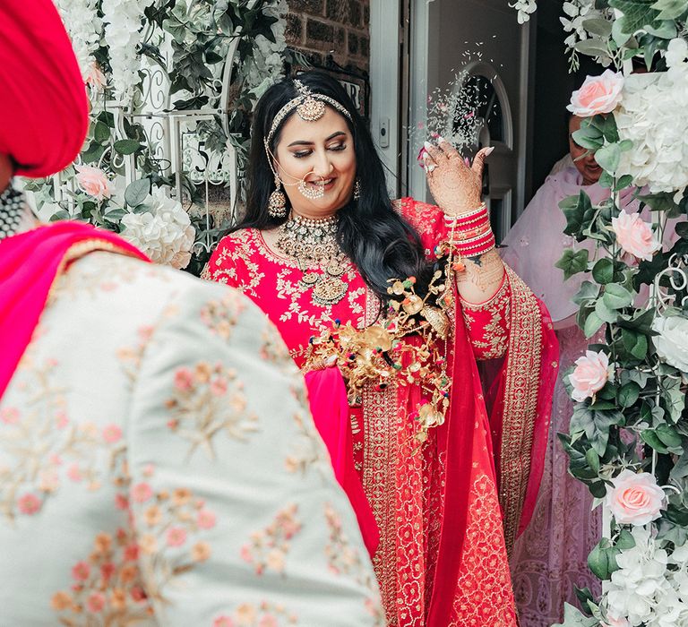 An Indian bride wears bright red traditional dress as she throws rice over shoulder.