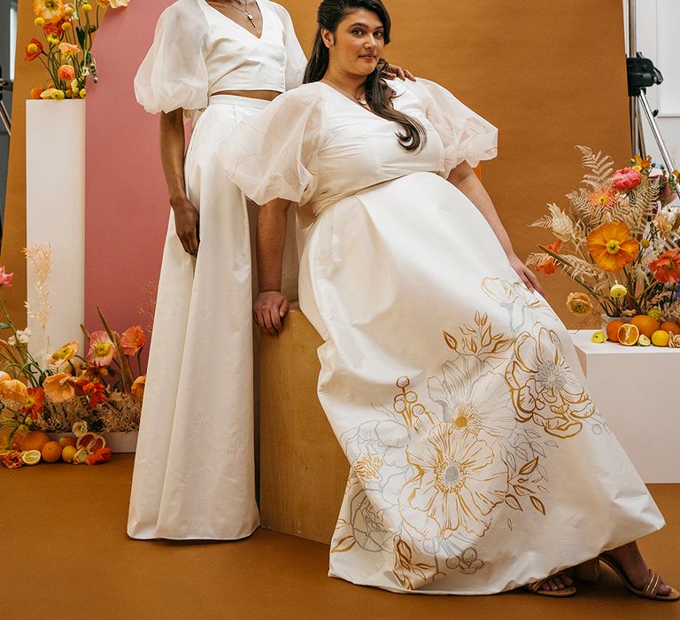 Two models wear bridal separates. Their outfits are white and they have gold detailing and jewellery.