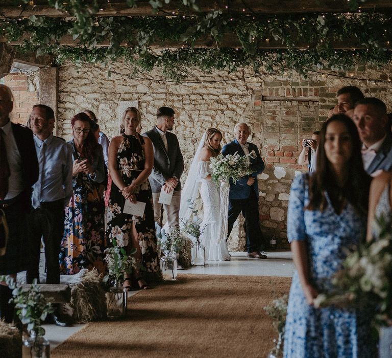 Bride in white Grace Loves Lace bardot wedding dress and veil holding white and green bridal bouquet walks into wedding ceremony with father in blue suit at Isle of Wight wedding with macrame wedding decor