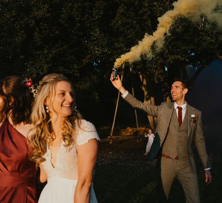 Groom in grey herringbone three piece suit with red tie and pocketsquare and floral buttonhole holds yellow smoke bomb next to bride in lace capped sleeve wedding dress during late summer wedding at Wellington Wood Norfolk