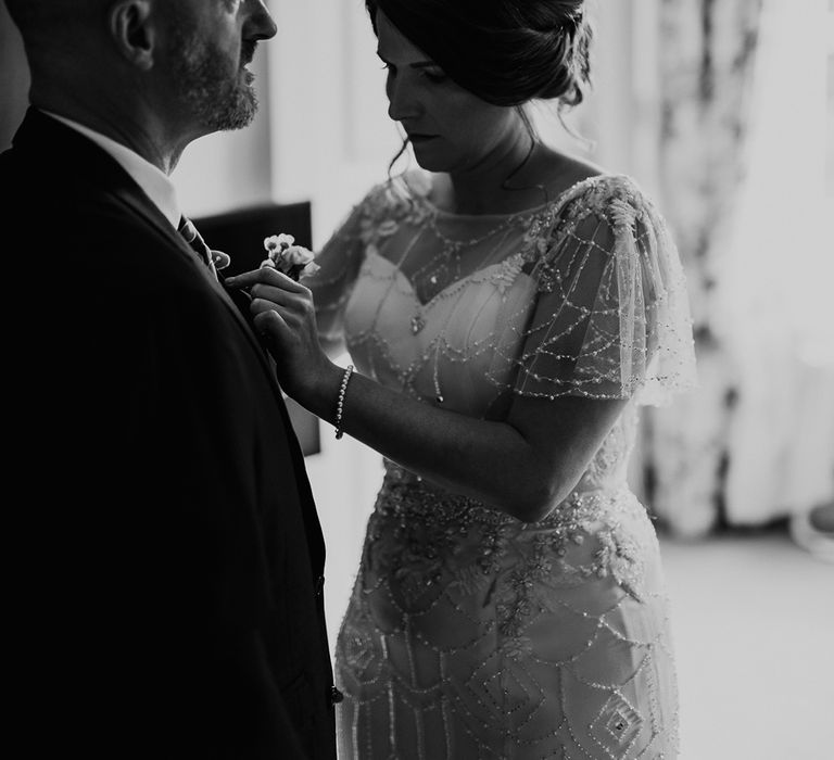 Bride in a vintage inspired wedding dress with embellishment putting on her fathers buttonhole flower 