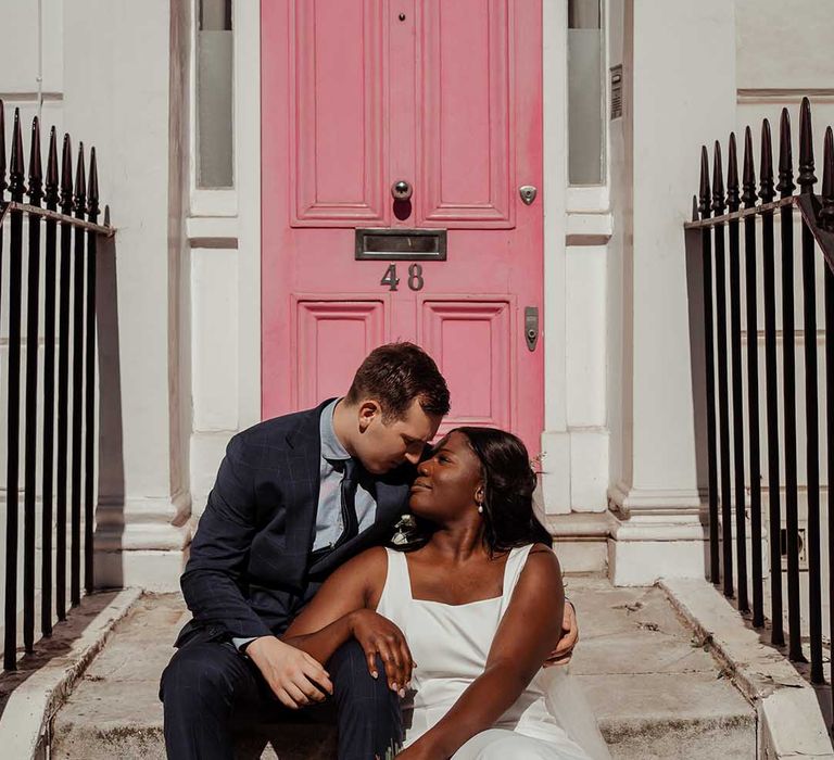 Bride & groom sit on steps outside pink door on their wedding day in Chelsea