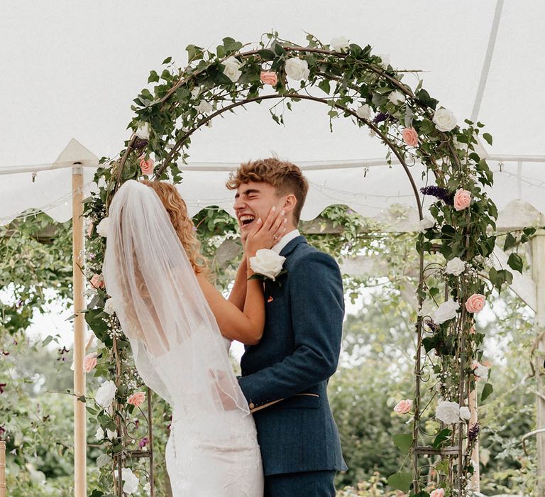 Bride & groom laugh and look lovingly one another during wedding ceremony whilst under floral archway