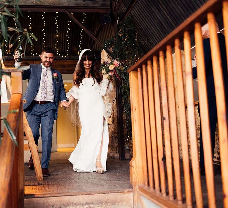 Bride in a wedding dress with front split showing off her gold Miu Miu platform shoes entering the wedding reception with her groom in a navy check suit 