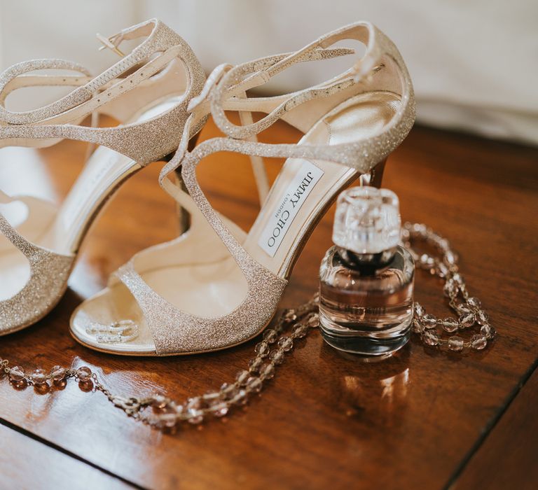 Gold sparkle strap Jimmy Choo wedding shoes with a bottle of perfume and rosary necklace on wooden table