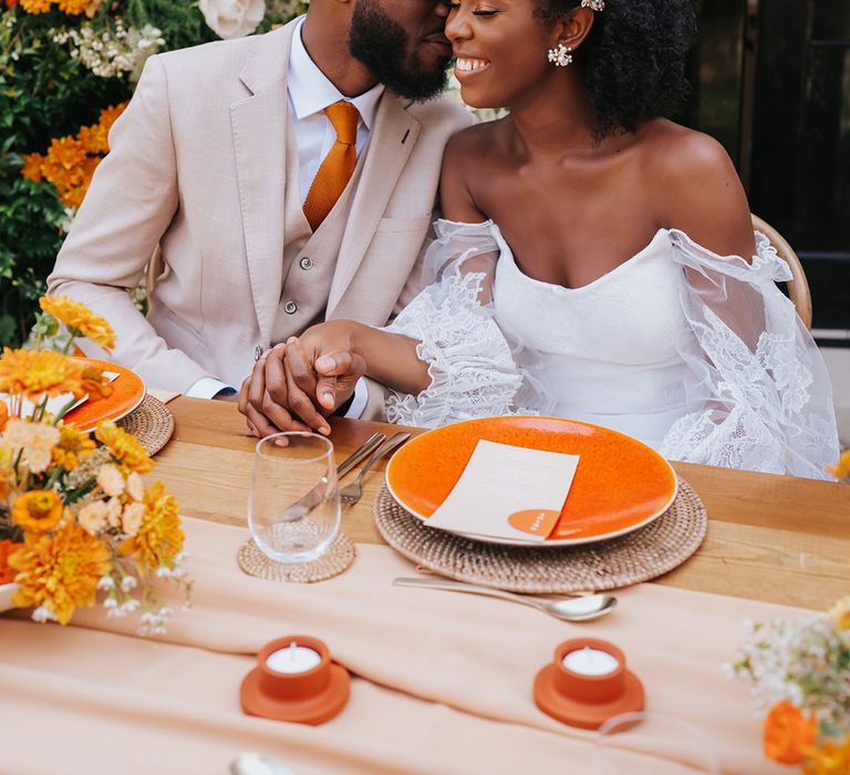 Groom in a beige suit and orange tie holding hands with his bride in a strapless wedding dress with lace sleeve at their orange outdoor wedding reception 