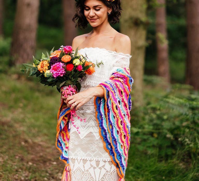 Smiling bride in off the shoulder Grace Loves Lace wedding dress and colourful flower crown holds colourful bridal bouquet and wears colourful crocheted blanket as she stands in woodland after outdoor wedding ceremony