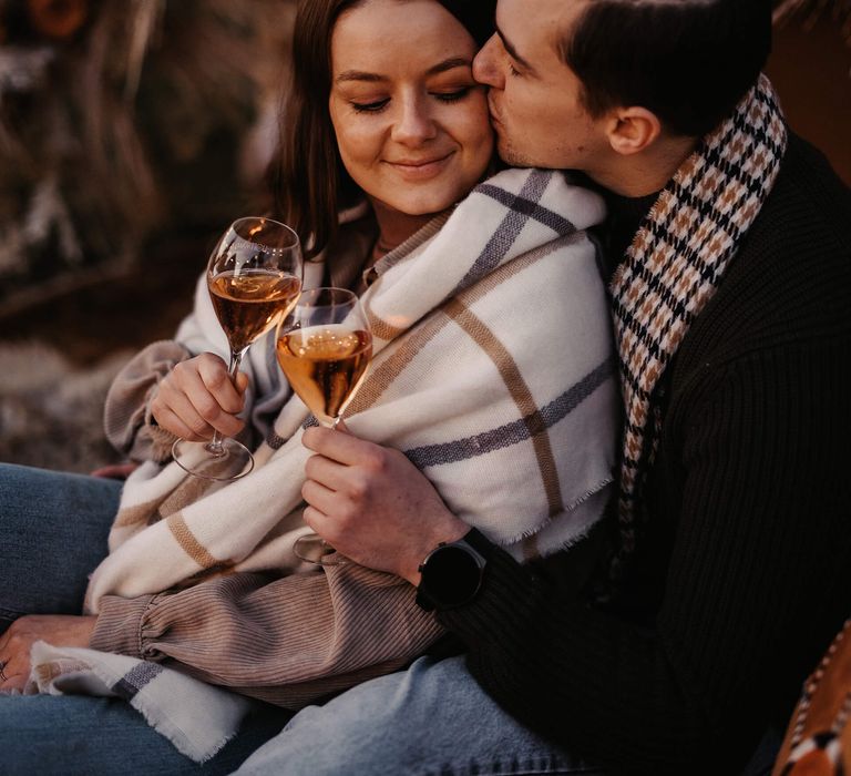 Groom-to-be kissing his fiancé's cheek at they snuggle at their intimate campsite proposal 