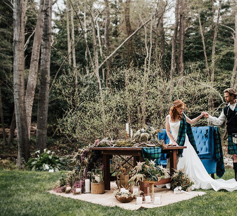 Groom in a blue and green wedding kilt holding hands with his bride in an A-line wedding dress at their outdoor boho sweetheart table reception 