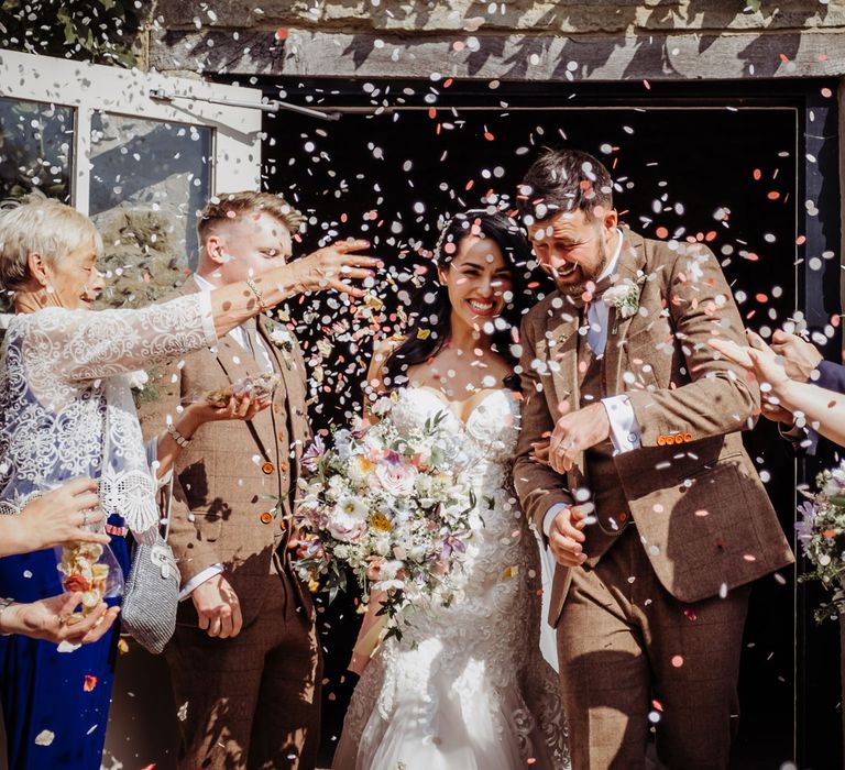 Bride in lace and tulle wedding dress with sweetheart neckline holding pastel bridal bouquet smiles whilst walking out of barn with groom in brown tweed suit as guests throw multicoloured confetti