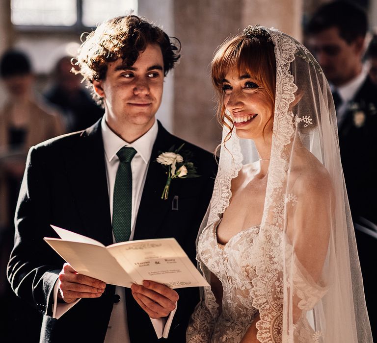 Groom gazes at bride as she beams during church ceremony in lace trim veil