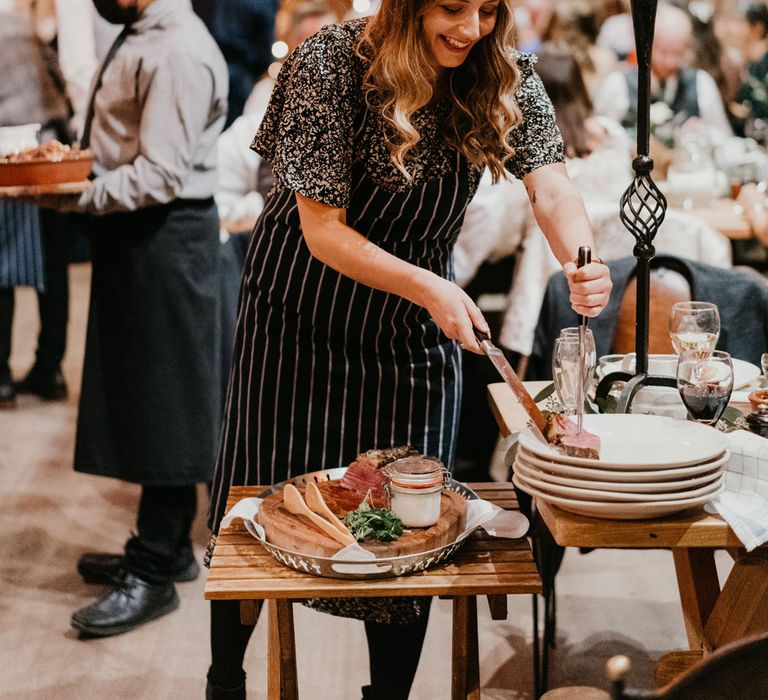 Guests in striped apron and chefs hat serves meat to a plate during barn wedding breakfast at The Tythe Barn in Launton