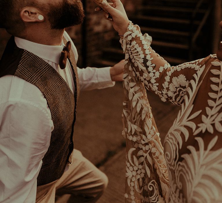 bride in along sleeve lace wedding dress feeding ice-cream to her groom in a tweed waistcoat and bow tie 