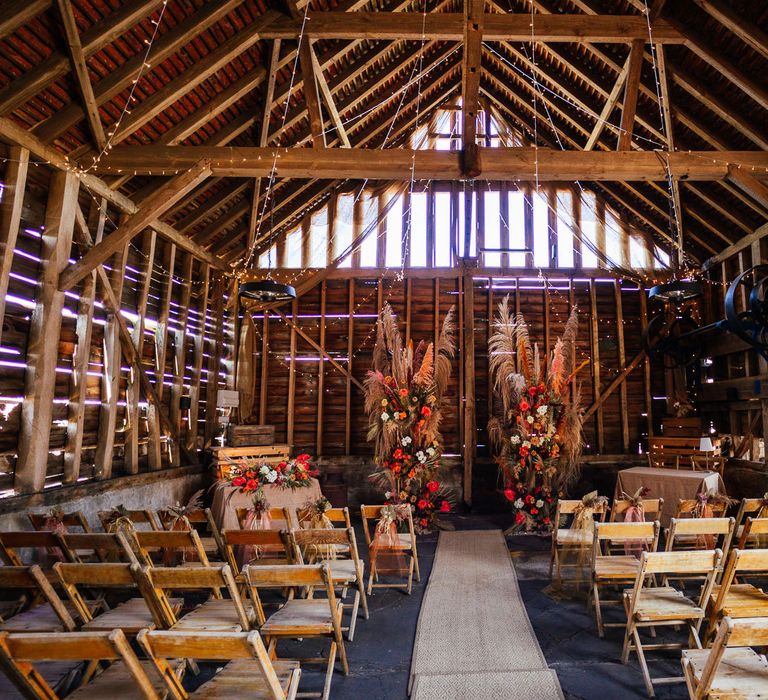 Barn wedding aisle with rustic orange and white pampas grass floral installations with wooden chairs and fairy lights