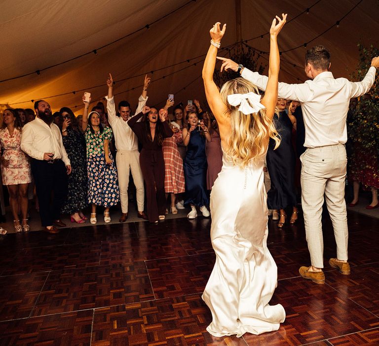 Bride in The Own Studio wedding dress with button detailing and satin hair bow dances with groom in white shirt and grey trousers on marquee dance floor