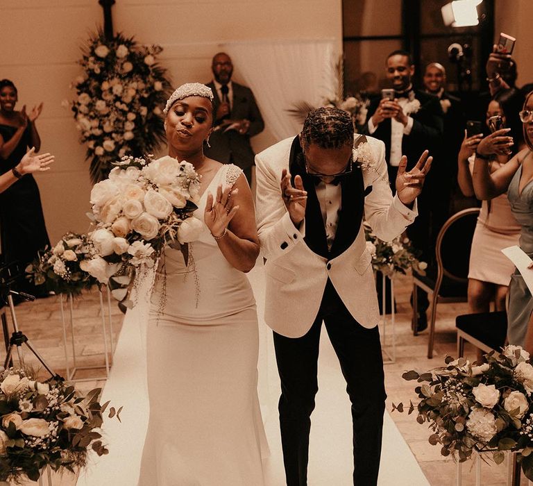 Black bride and groom in a vintage style wedding dress and beaded headdress and white tuxedo jacket dancing down the aisle 