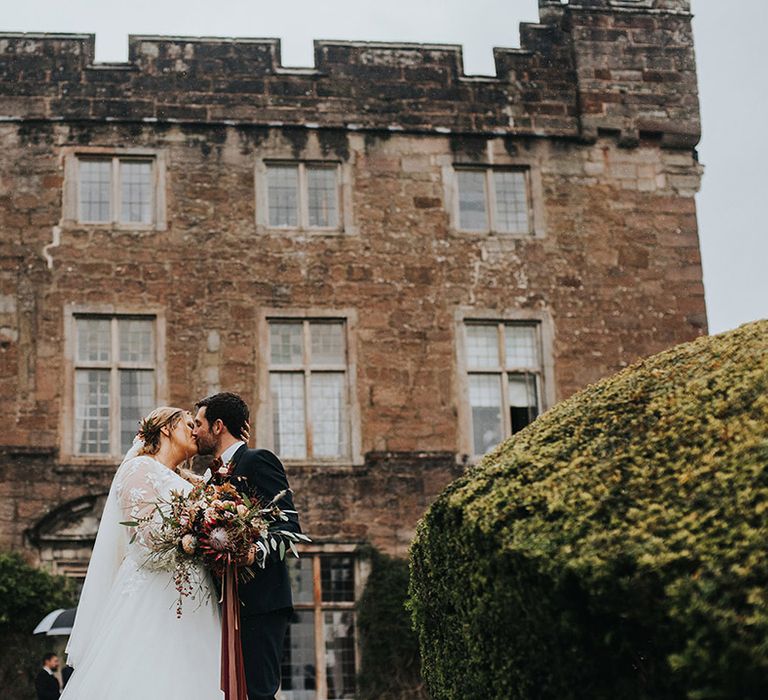 Bride in a long sleeve wedding dress holding a Burgundy protea wedding bouquet as she kissed her groom in a navy suit on the grounds of Askham Hall