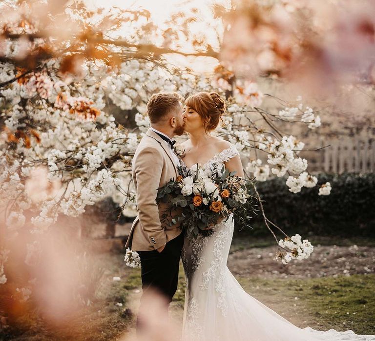 Bride and groom share a kiss under the blossom trees for spring wedding in rustic barn venue