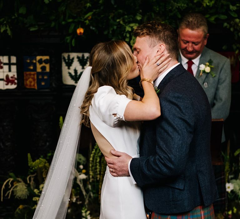 Bride in cutout back puff sleeve wedding dress with pearl veil kisses groom in navy blue suit and tie with kilt