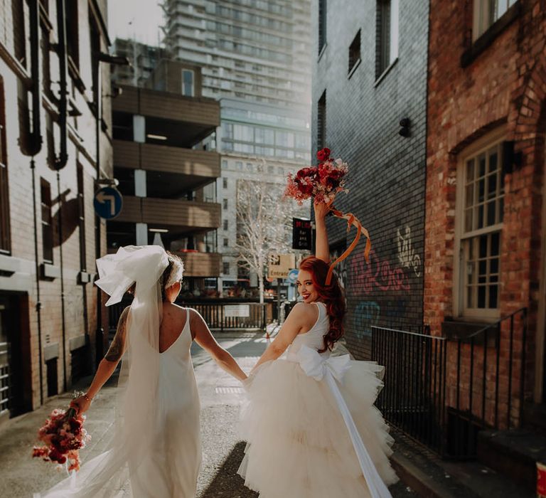 Manchester elopement with two brides in a layererd tulle wedding dress holding a red bouquet in the air and a satin slip wedding dress long bow veil 