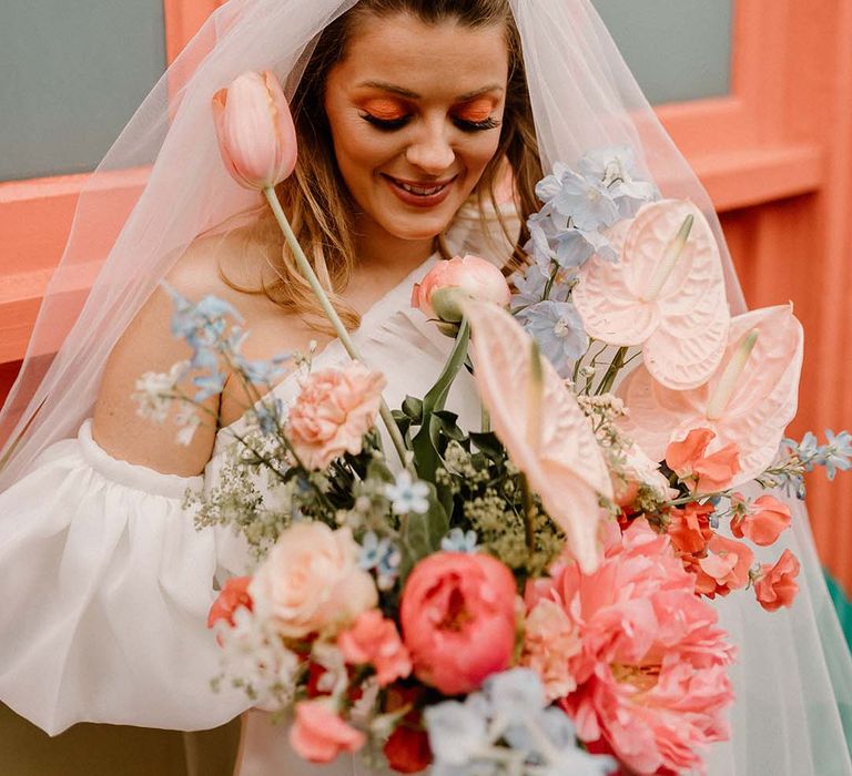 Bride with coral eye shadow wedding makeup and false eyelashes holding a pink and blue wedding bouquet with anthuriums, peonies and delphiniums 