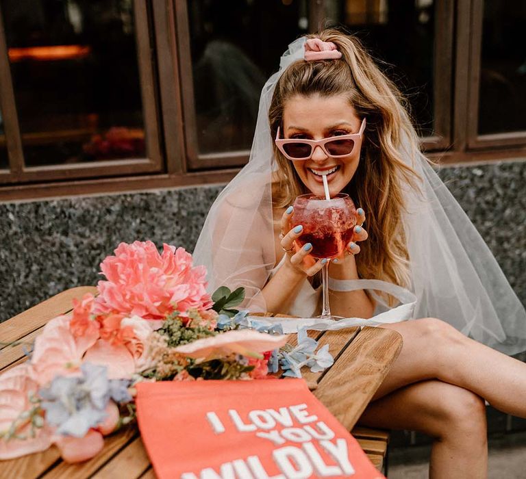 Bride in retro pink sunglasses and a short wedding dress drinking a cocktail with a pink suede fabric wedding banner on the table 