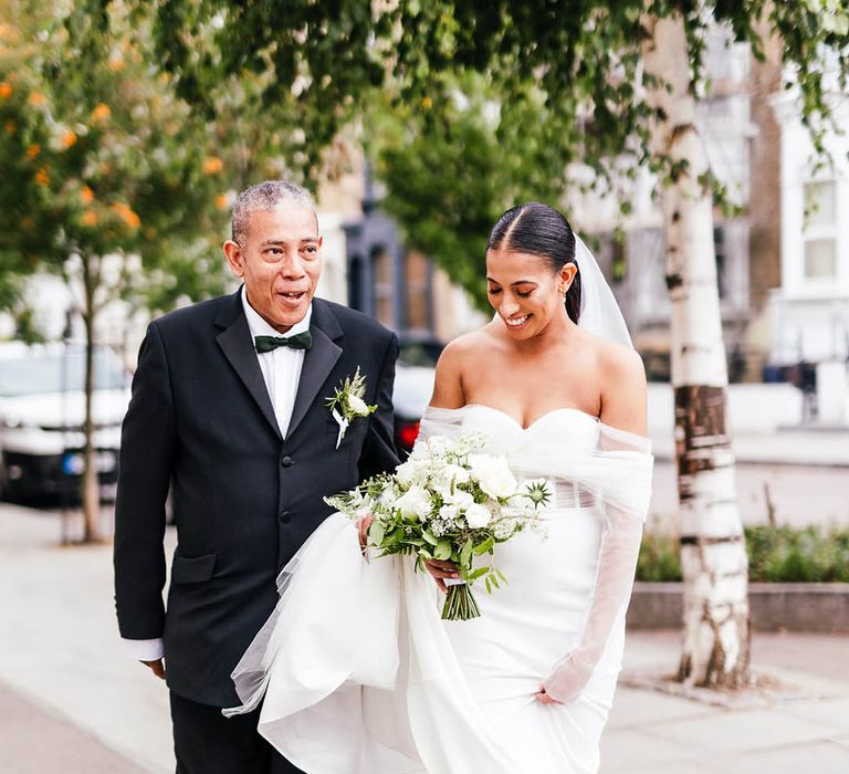 Father of the bride in black tie walks with bride in off the shoulder corset top wedding dress