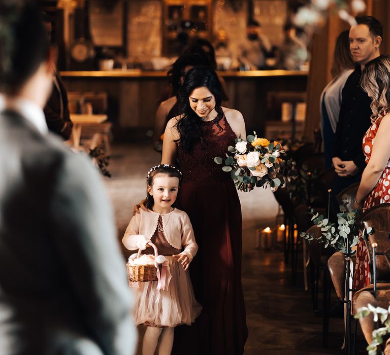 Flower girl in pale pink dress holding wicker basket of flower petals walks with bridesmaid in deep red burgundy dress with sparkles 