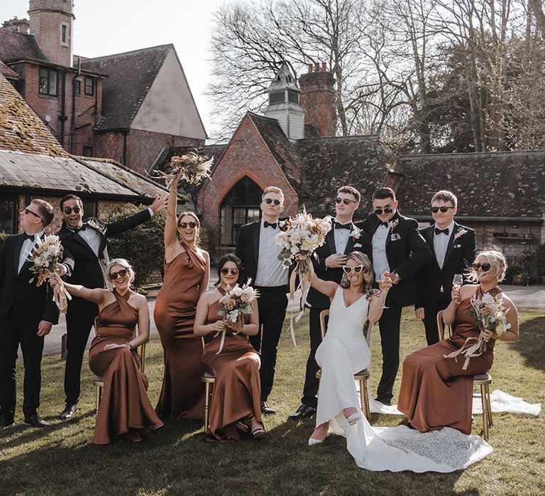 Groom and groomsmen in black tie and bride with bridesmaids in copper dresses all wearing sunglasses on sunny wedding day 