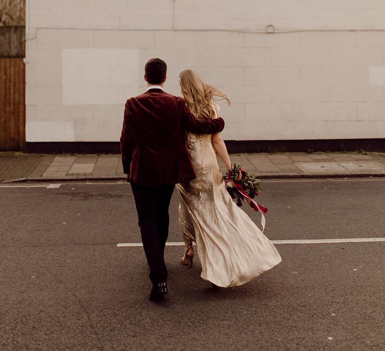 Groom in velvet suit jacket holds the bride in sparkly gold wedding dress walk around their city wedding venue together 