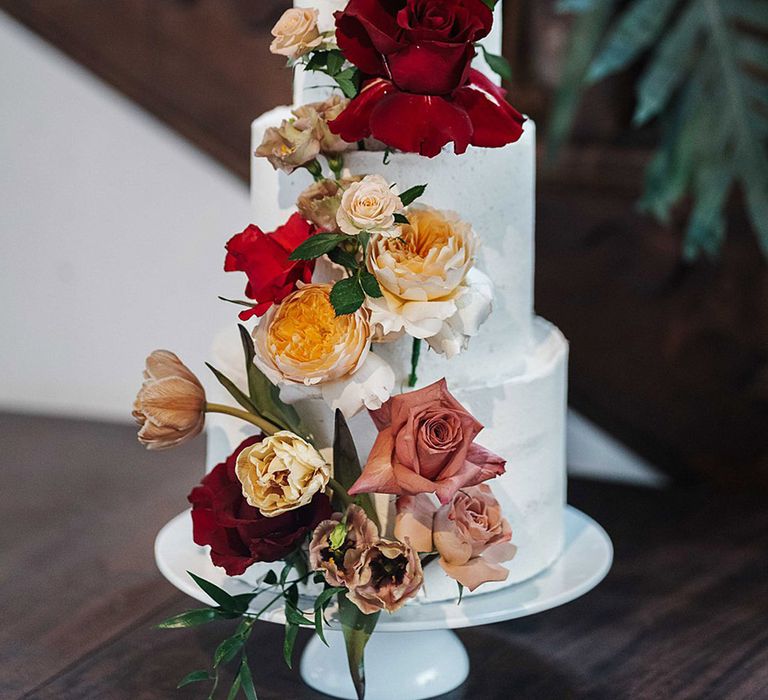 Three tier wedding cake frosted with white icing and decorated with different coloured roses including red, toffee and pink ones