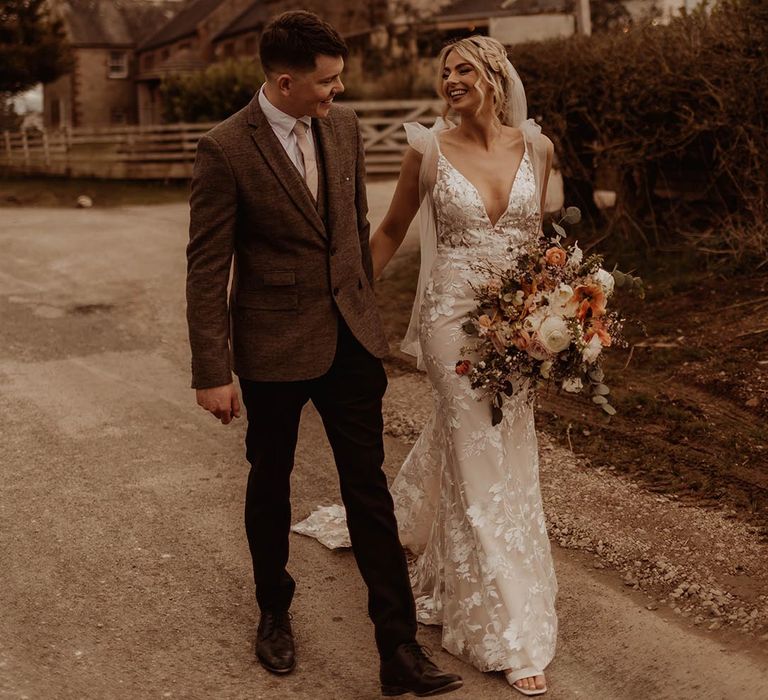 Bride in v-neck lace wedding dress with bow sleeves walking with the groom in brown suit holding orange and white bouquet